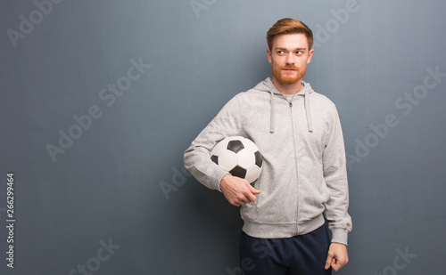 Young redhead fitness man smiling confident and crossing arms, looking up. He is holding a soccer ball.