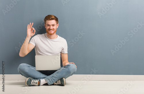 Young redhead student man sitting on the floor cheerful and confident doing ok gesture. He is holding a laptop.
