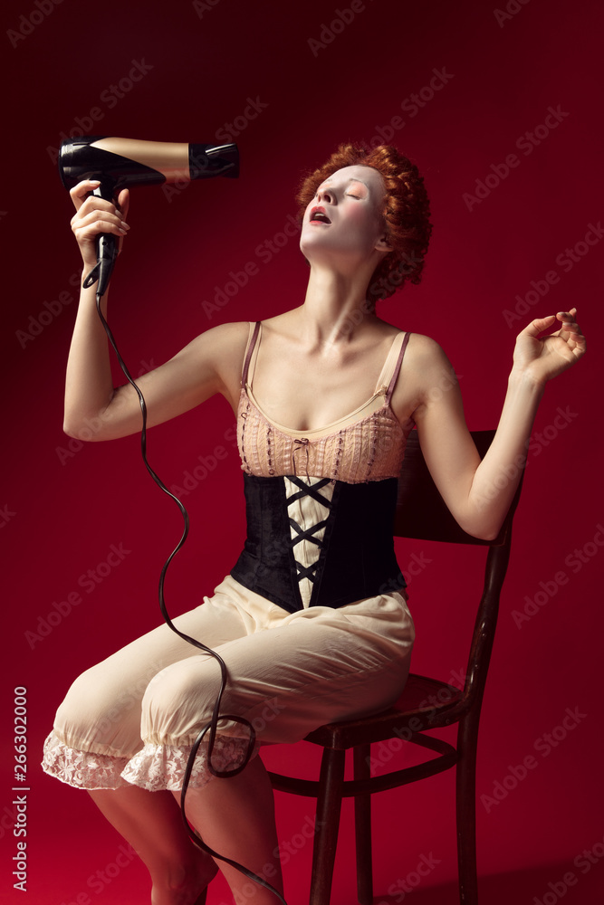 Medieval redhead young woman as a duchess in black corset and night clothes sitting on the chair on red background. Doing her hair with dryer. Concept of comparison of eras, modernity and renaissance. Medieval redhead young woman as a duchess in black corset and night clothes sitting on the chair on red background. Doing her hair with dryer. Concept of comparison of eras, modernity and renaissance.
