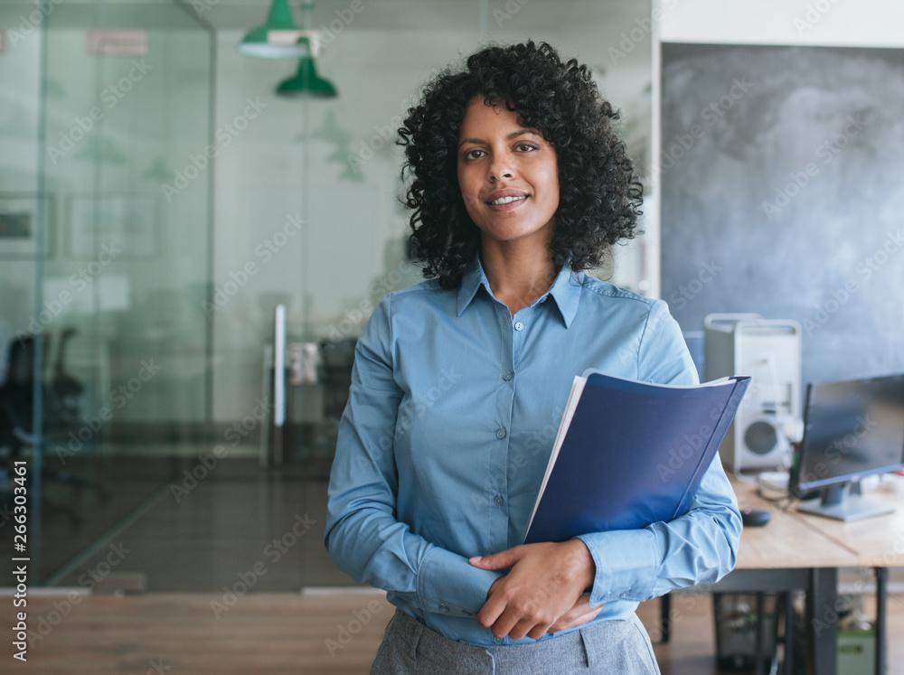 © mavoimages - Smiling young businesswoman carrying paperwork while standing in an office © mavoimages - Smiling young businesswoman carrying paperwork while standing in an office