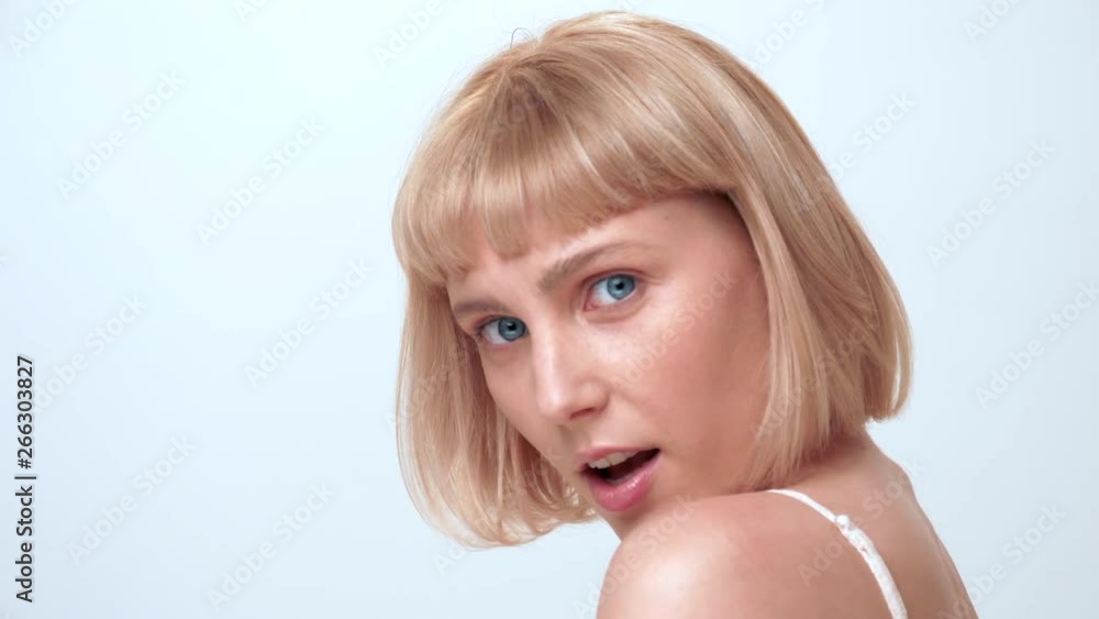 A close-up portrait of a blonde young caucasian woman with blue eyes turning around and surprisingly looking into the camera for a beauty shoot in the studio on a white background.