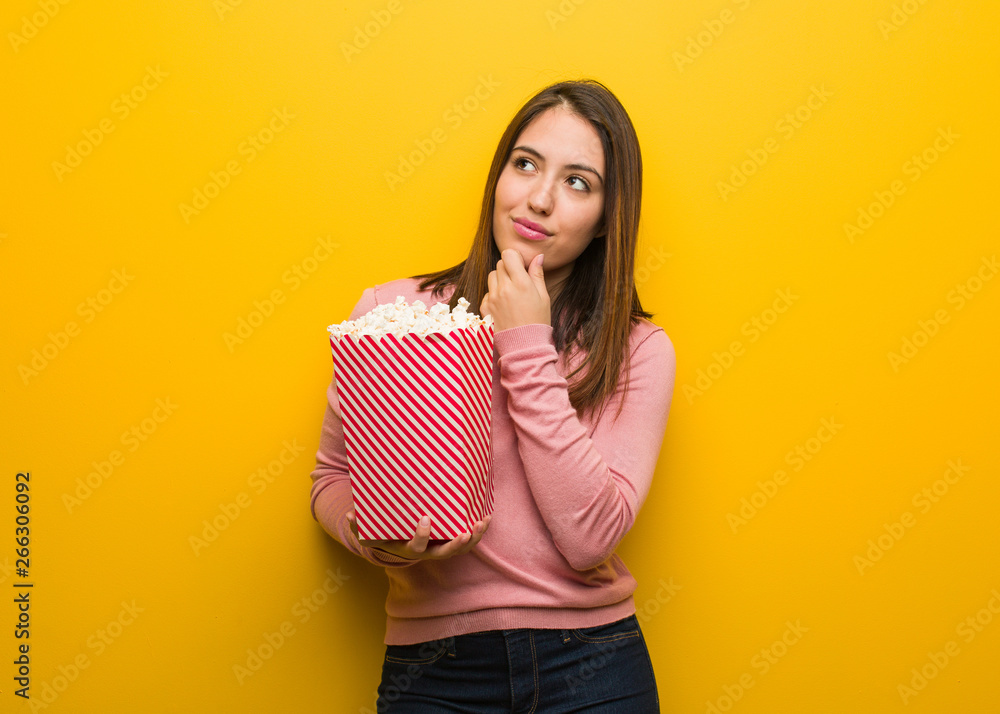 Young cute woman holding a popcorn bucket doubting and confused Young cute woman holding a popcorn bucket doubting and confused