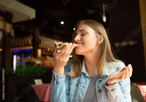 Funny blonde girl in jeans jacket eating pizza at restaurant. 