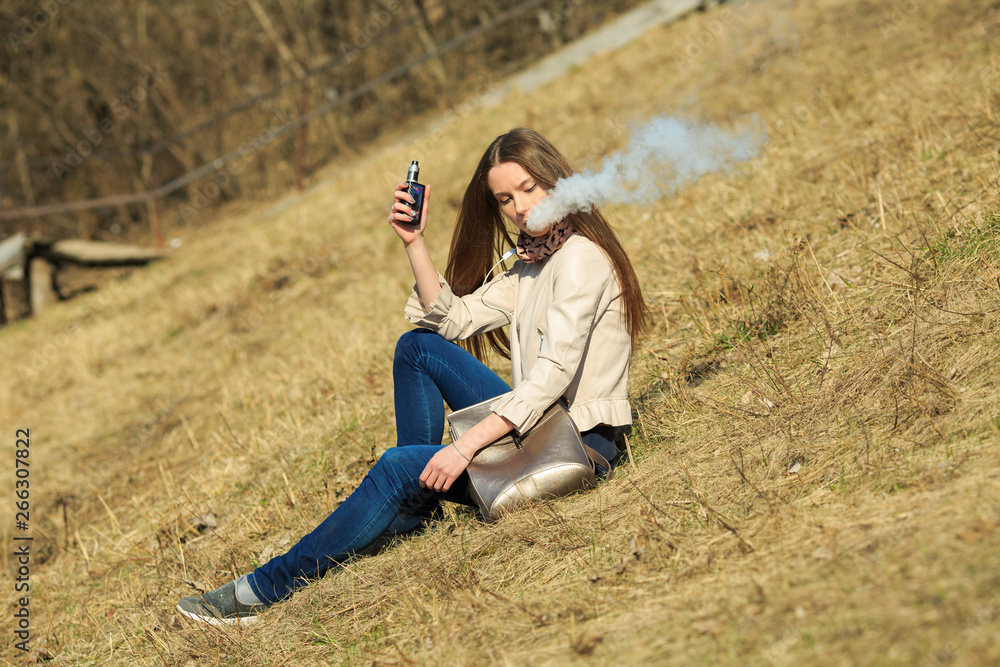 Vape teenager. Young pretty white girl in casual clothing smoking an electronic cigarette on the hillside on a sunny day in the spring. Bad habit. Vaping activity. Vape teenager. Young pretty white girl in casual clothing smoking an electronic cigarette on the hillside on a sunny day in the spring. Bad habit. Vaping activity.