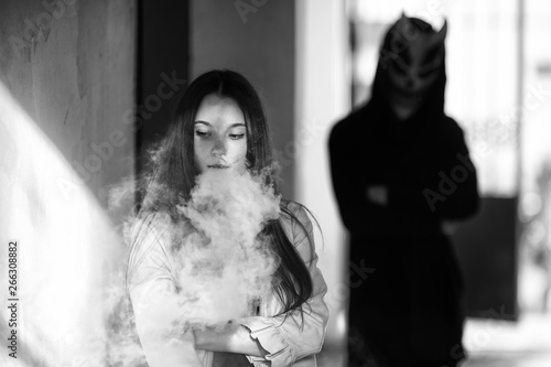 Vape teenager and death. Young cute girl in a dress an electronic cigarette near the wall in front of monster in the background outdoors in spring. Vaping activity. Black and white.