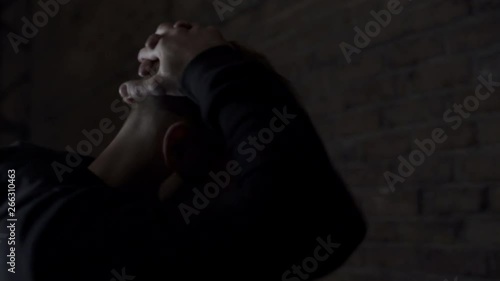 Shoulder camera shot in 4K of a angry handsome blond young man in hooded sweater banging his head with his fists with a light moving in a studio shed brick wall