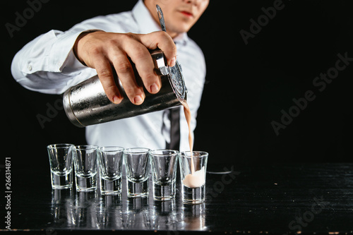Barman makes white cocktail shots out of shaker. Shooter making shots at a bar. Night out with white alcohol shots on the table. 