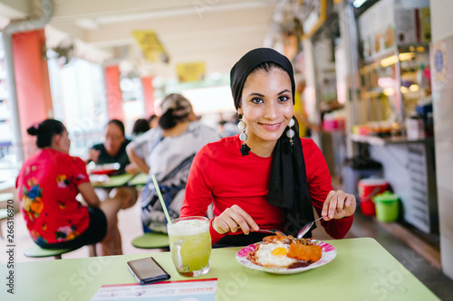 Canvas Print A beautiful and attractive Malay Muslim woman in a hijab enjoys nasi lemak and sugarcane juice for lunch during the daytime in a hawker center in Singapore