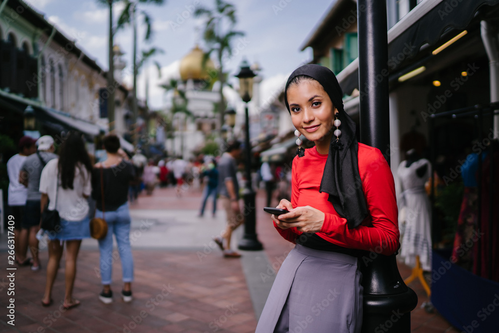 A beautiful and young Muslim Middle Eastern woman texts on her smartphone as she leans against a pole on a crowded street with a mosque in the distance. She is smiling confidently. A beautiful and young Muslim Middle Eastern woman texts on her smartphone as she leans against a pole on a crowded street with a mosque in the distance. She is smiling confidently.