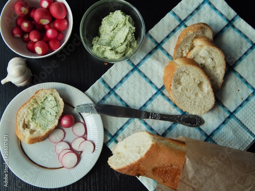 Fresh white baguette and a glass bowl with green butter made of butter, dill, garlic and salt and white bowl with organic radish, slice of baguette with green butter and sliced radish. Healthy food.