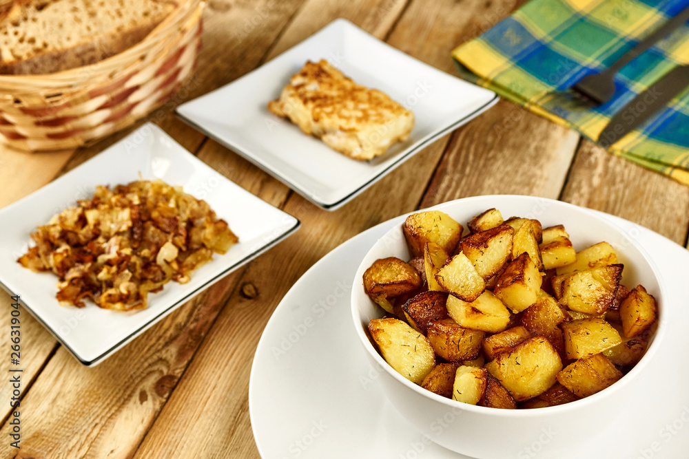 Roasted potatoes in a white bowl with rosemary and dill at next to fried onions and fish in batter on wooden background. Close-up rustic composition. Roasted potatoes in a white bowl with rosemary and dill at next to fried onions and fish in batter on wooden background. Close-up rustic composition.
