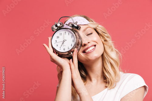 Portrait of beautiful young woman 20s wearing sleeping mask holding alarm clock after awakening