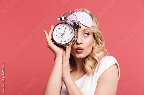 Portrait of charming young woman 20s wearing sleeping mask holding alarm clock after awakening