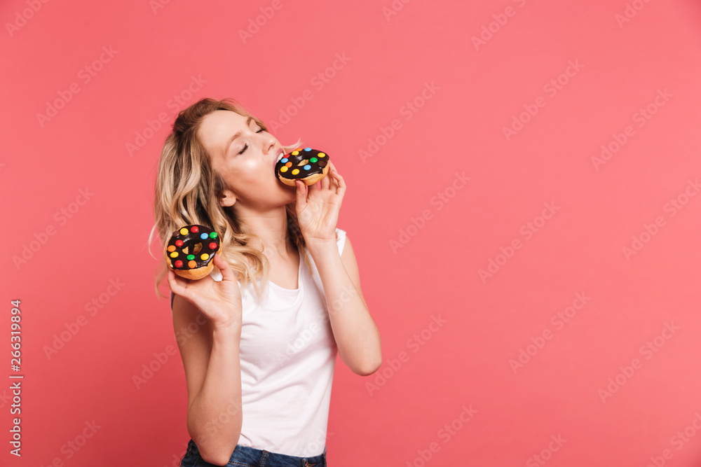 Portrait of happy blond woman 20s wearing casual t-shirt holding and eating tasty sweet donuts Portrait of happy blond woman 20s wearing casual t-shirt holding and eating tasty sweet donuts