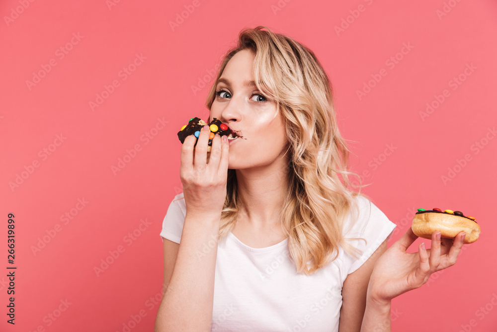 Portrait of european blond woman 20s wearing casual t-shirt holding and eating tasty sweet donuts