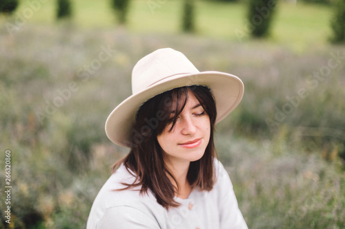 Stylish hipster girl in linen dress and hat sitting on bench at lavender field and relaxing in the morning. Portrait of bohemian woman enjoying vacation in mountains. Atmospheric moment