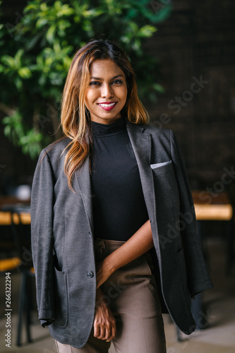 Mid shot Portrait of a tanned, professional young Southeast Asian business woman in a dark turtle neck blouse with blazer and slacks standing in the city restaurant and is smiling during the day.