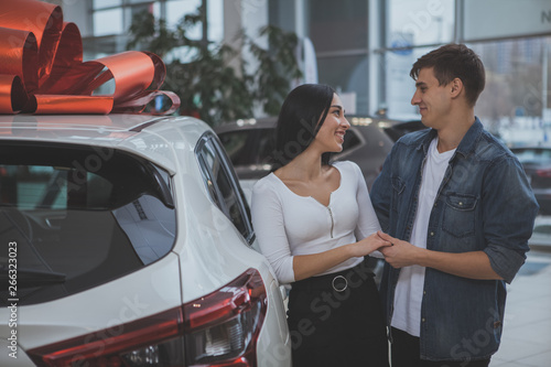 Happy young couple holding hands smiling at each other while choosing new car at the dealership salon. Cheerful beautful woman and her husband buying new auto. Transportation vehicle concept