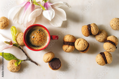 Baci di dama homemade italian hazelnut biscuits cookies with chocolate cream served with red cup of espresso coffee and magnolia flowers over white marble background. Flat lay  space
