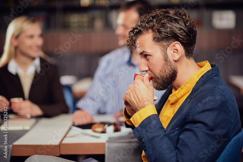Bearded businessman dressed smart casual drinking coffee while sitting in cafe. In background his colleagues discussing about project.