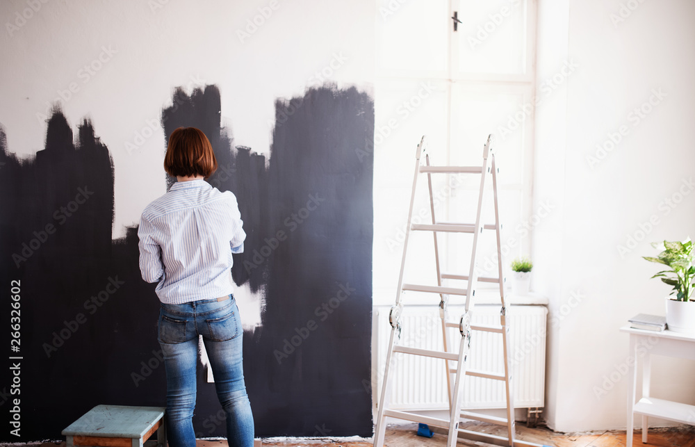 A rear view of young woman painting wall black. A startup of small business.