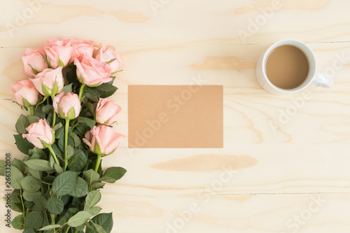 Top view of a kraft card mockup with a bouquet of pink roses and a coffee on a wooden table.