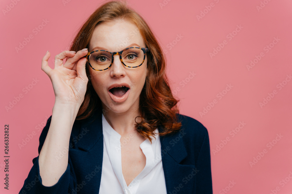 Headshot of surprised red haired woman keeps hand on frame of glasses, jaw dropped, can not believe her eyes, wears elegant suit, shocked by unexpected rumor, stands over pink studio wall, free space Headshot of surprised red haired woman keeps hand on frame of glasses, jaw dropped, can not believe her eyes, wears elegant suit, shocked by unexpected rumor, stands over pink studio wall, free space