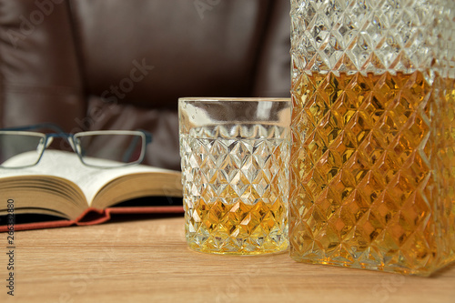 Alcoholic drink in crystal decanter. In a glass poured whiskey on a wooden table in the background a chair for seating. Open book with glasses.