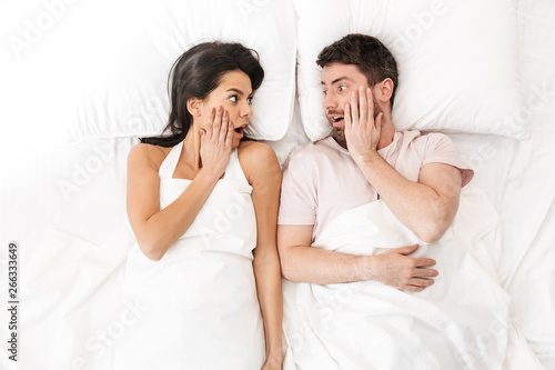 Shocked loving couple lies in bed under blanket.