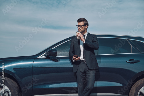 Moment to relax. Handsome young businessman using smart phone and drinking coffee while standing near his car outdoors © gstockstudio