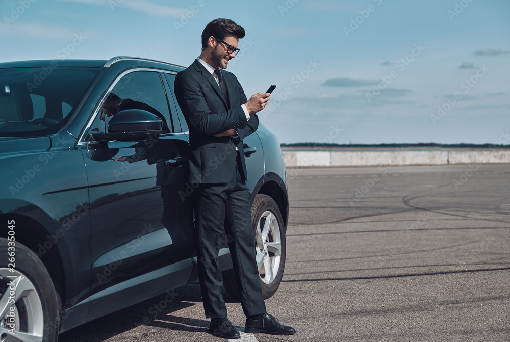 Confident manager. Full length of handsome young businessman using smart phone while standing near his car outdoors