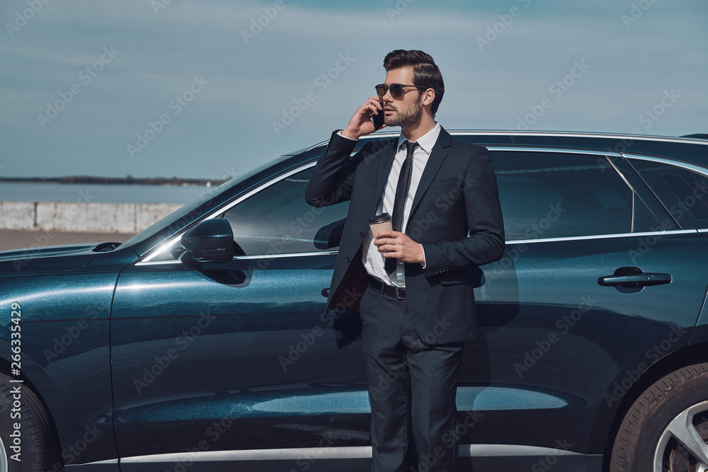 Time is money. Handsome young businessman talking on his smart phone and smiling while standing near his car outdoors Time is money. Handsome young businessman talking on his smart phone and smiling while standing near his car outdoors