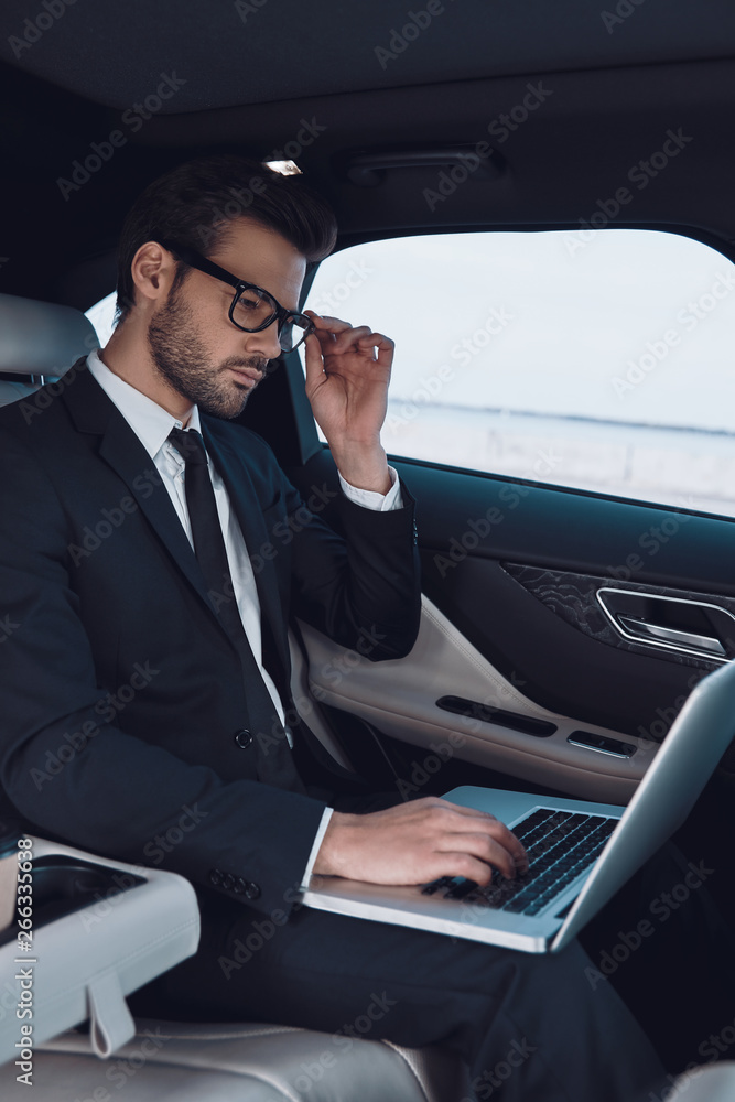 Business is his life. Thoughtful young man in full suit working using laptop and adjusting his eyewear while sitting in the car