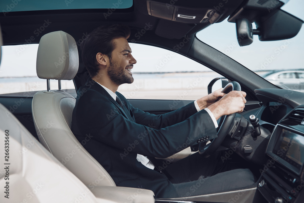 In love with high speed. Handsome young man in full suit smiling while driving a car