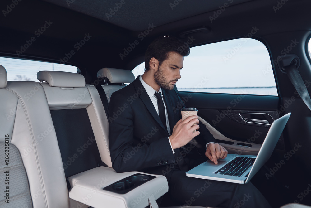 Business on the go. Handsome young man in full suit working using laptop while sitting in the car