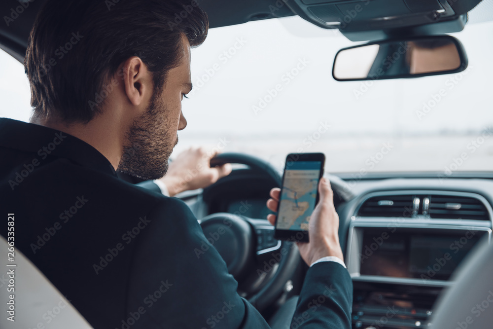 Where am I? Rear view of young man in full suit using smart phone to check the map while driving a car Where am I? Rear view of young man in full suit using smart phone to check the map while driving a car