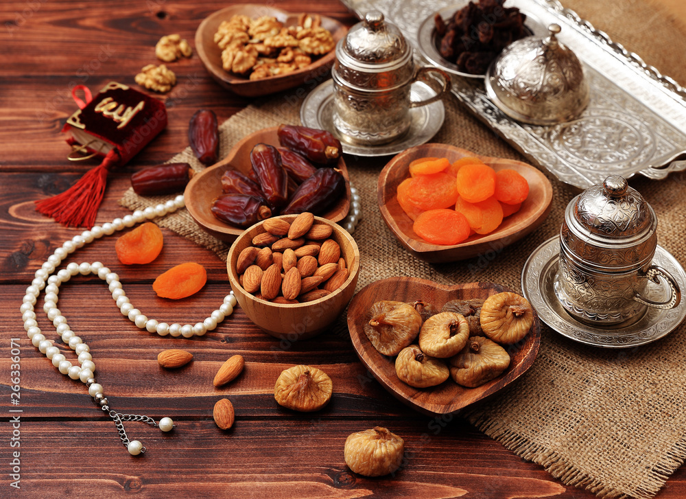 Ramadan concept. Delicious dates, dried figs, dried apricots, walnuts, almonds, raisins on bamboo and silver plates, rosary beads with Islamic holy book Quran on a pine wooden table background.