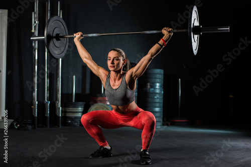 Young, European, muscular girl in red leggings, doing exercise with a barbell in the gym for crossfit. Dark background