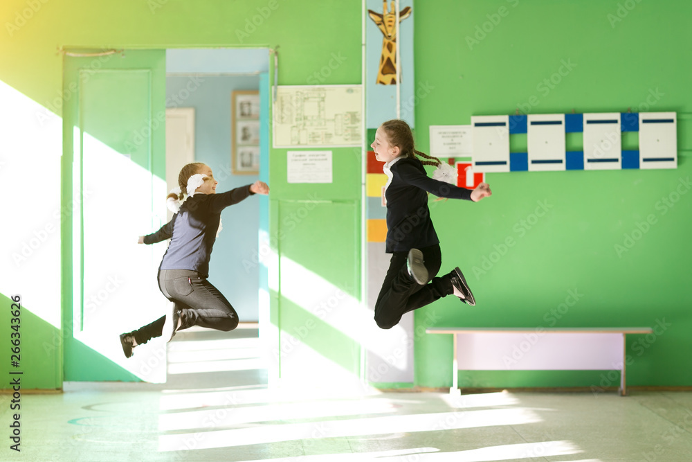 Fototapeta premium Cheerful children jump in the school hallway. Two girls schoolgirls jump in height at school during holidays.