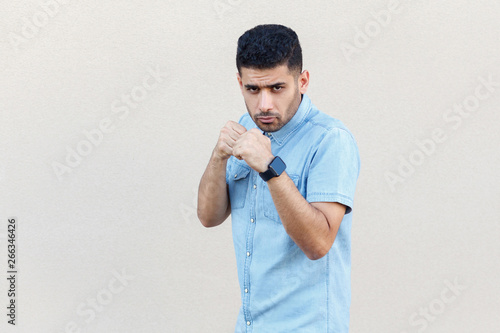 Portrait of serious handsome young bearded businessman in blue shirt standing with boxing fists and ready to attack and looking at camera. indoor studio shot isolated on light beige wall background.