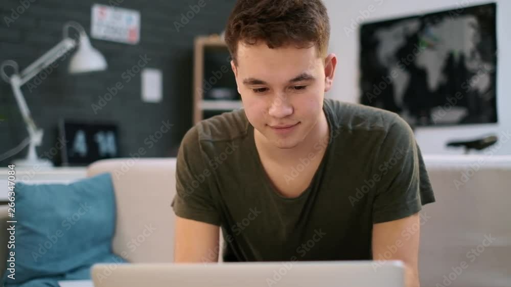 A teenage caucasian boy is happy working on the laptop in his room and nods his head A teenage caucasian boy is happy working on the laptop in his room and nods his head