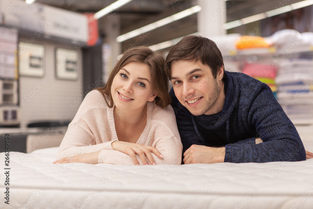 Attractive young woman and her cheerful handsome boyfriend embracing, lying on the bed together at home furnishings store. Happy couple buying orthopedic mattress and bed for their new home Attractive young woman and her cheerful handsome boyfriend embracing, lying on the bed together at home furnishings store. Happy couple buying orthopedic mattress and bed for their new home