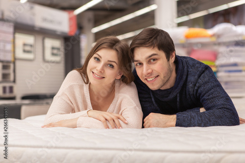 Attractive young woman and her cheerful handsome boyfriend embracing, lying on the bed together at home furnishings store. Happy couple buying orthopedic mattress and bed for their new home