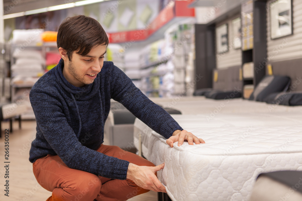 Handsome cheerful man examining orthopedic mattress at furniture store, copy space. Young man shopping at home furnishings department, choosing new bed to buy. Rest, relaxation, health concept Handsome cheerful man examining orthopedic mattress at furniture store, copy space. Young man shopping at home furnishings department, choosing new bed to buy. Rest, relaxation, health concept