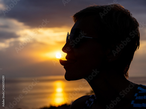 Silhouette of a woman at sunset close-up, playfully showing the tip of the tongue, trying to touch the outgoing sun on the background of the sea landscape