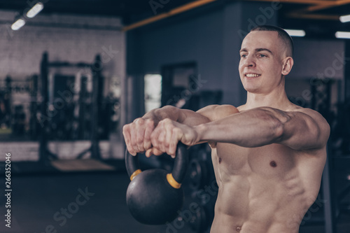 Happy handsome shirtless fitnessman enjoying working out with kettlebell at crossfit box gym, copy space. Cheerful healthy sportsman smiling, lifting weights at sports studio. Motivation, lifestyle co photo