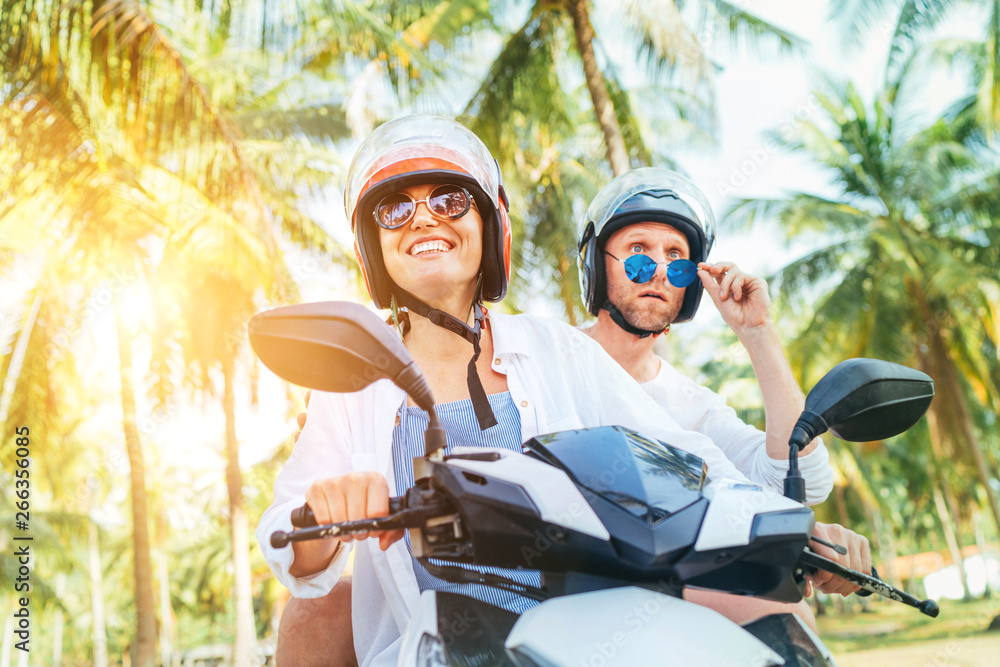 Сouple travelers riding motorbike scooter in safety helmets during tropical vacation under palm trees - obrazy, fototapety, plakaty 