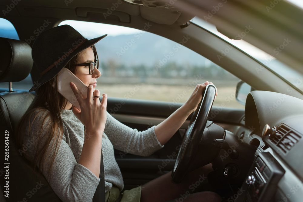 Young hipster woman with eyeglasses and hat driving the car and holding smartphone