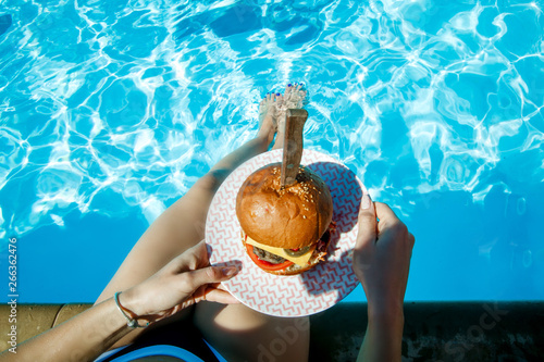 Top view of juicy Burger in girl's hands and feet in the pool. Concept burgers
