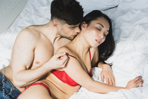 handsome shirtless man kissing passionate asian girlfriend in red lingerie